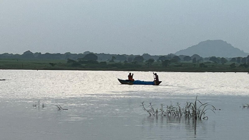A small boat carrying two people moves across a calm lake, with grassy shoreline, scattered trees, and a hazy hill in the background.”