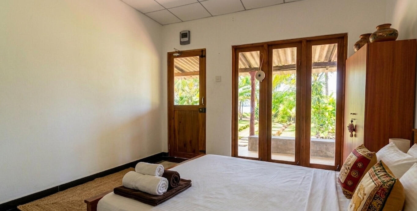 Spacious bedroom with wooden glass doors, natural sunlight, portable air conditioners, and a peaceful garden view."